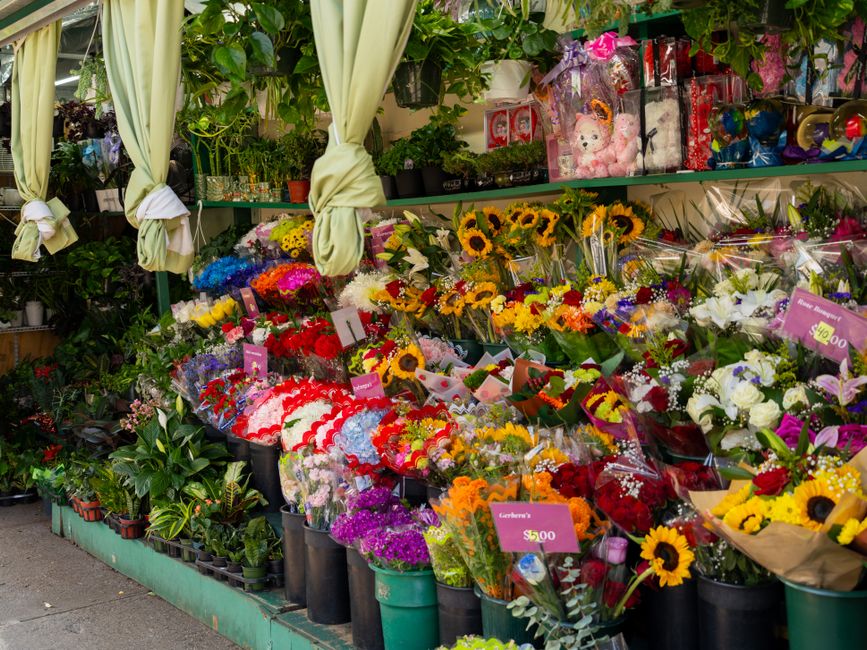 An outdoor display of fresh floral bouquets arranged on a green stand and shelves at a flower shop. The arrangement includes sunflowers, roses, white lilies, and various mixed flowers in vibrant color