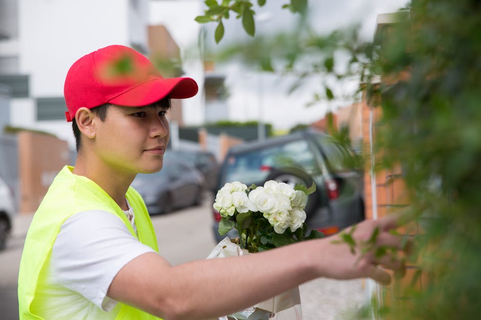 A delivery person wearing a red cap and a high-visibility yellow vest is placing a fresh floral gift of white hydrangeas into a green plant holder outside a residential building. The arrangement featu
