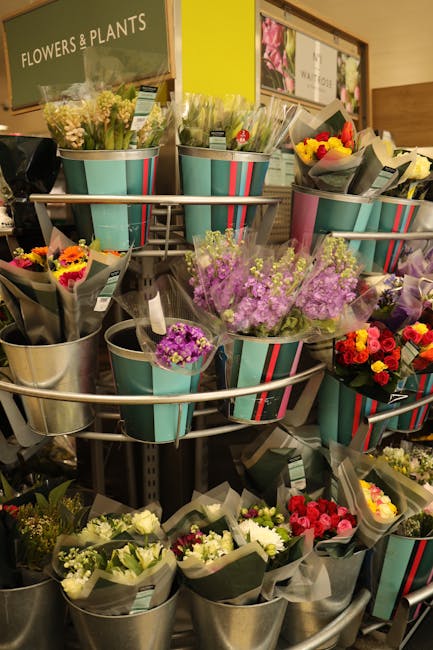 A display of various fresh flowers arranged in metal buckets on a tiered stand inside a florist shop, with a green sign reading 'FLOWERS & PLANTS' in the background. The bouquets feature a range of co