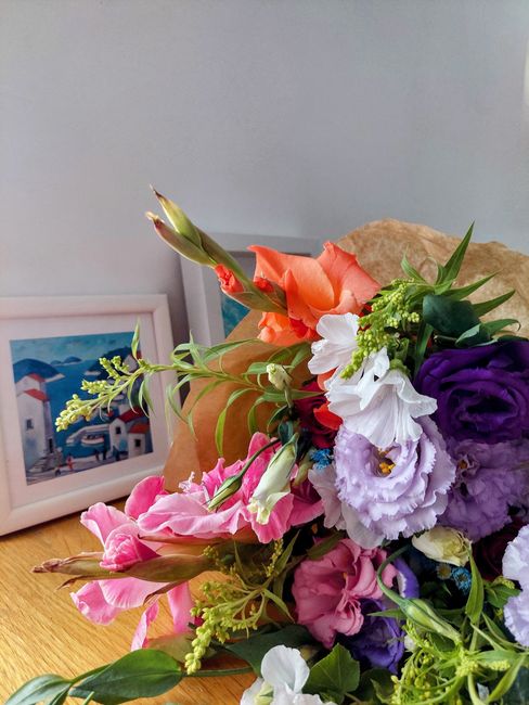A vibrant bouquet of fresh flowers resting on a wooden surface, featuring pink and white snapdragons, orange gladiolas, purple lisianthus, white eustomas, and green foliage, wrapped in brown paper wit